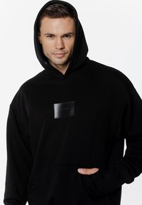 Czarna bluza z kapturem, z przednią kieszenią i prostok ątną skórzaną naszywką z logo. Miękka tkanina, luźny krój i długie rękawy.