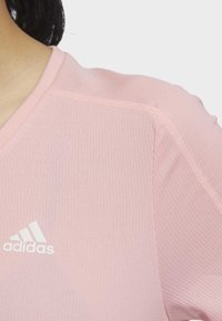 Ljust rosa Adidas topp med v-ringning, texturerat tyg och vit logotyp på vänster bröst, med synliga sömnadsdetaljer.