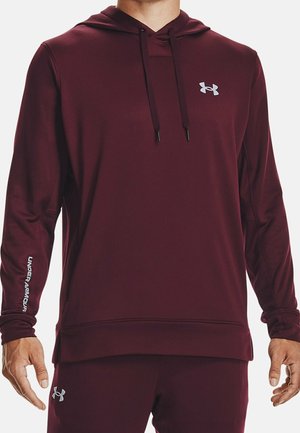 Mężczyzna ubrany w bordową bluzę z kapturem Under Armour oraz dopasowane spodnie, obie z widocznymi logo na piersi, rękawie i udzie.