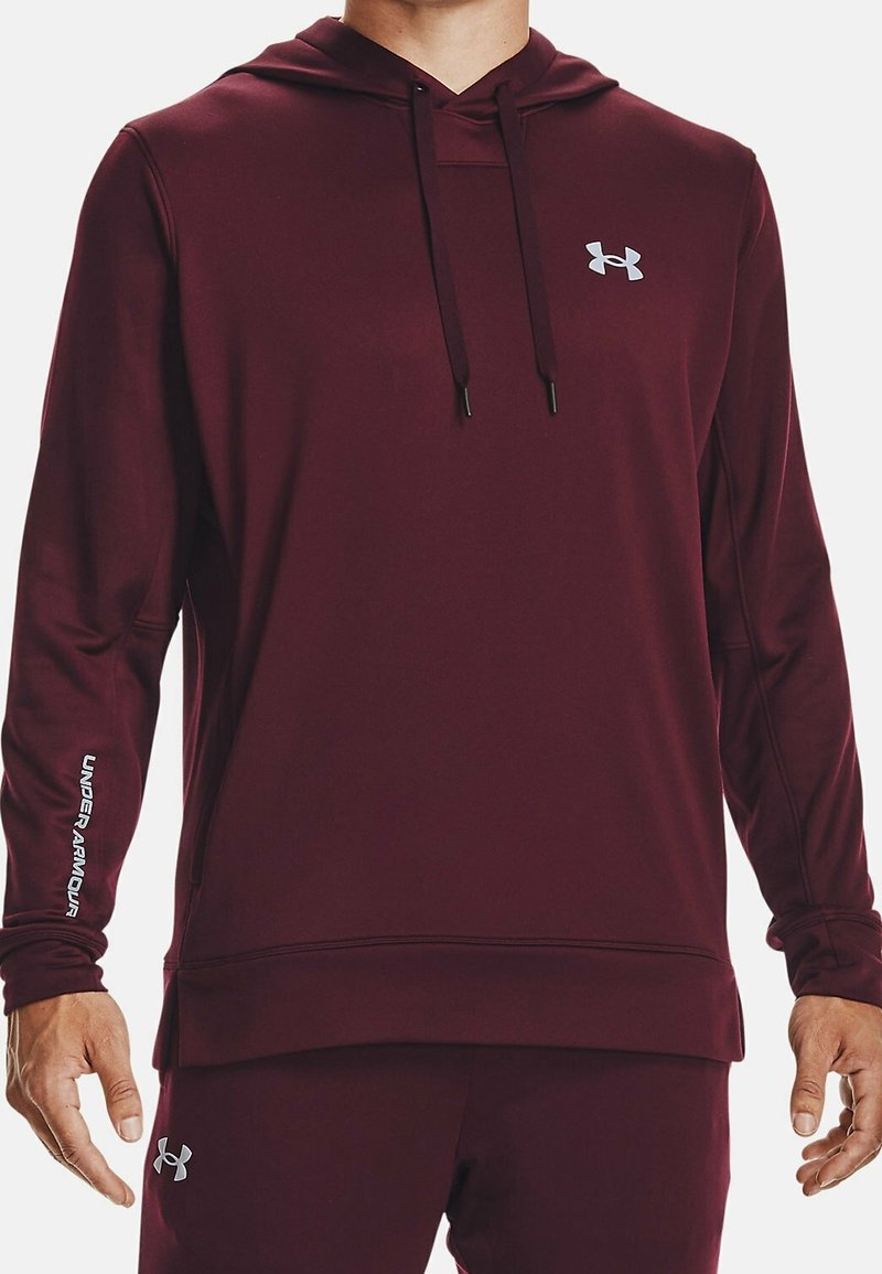 Homme portant un sweat à capuche Under Armour bordeaux et un pantalon assorti, tous deux avec des logos visibles sur la poitrine, la manche et la cuisse.