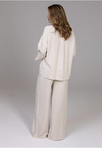Mujer con pelo largo y ondulado, vistiendo un suéter de punto suelto color beige claro y pantalones anchos a juego, de pie frente a un fondo gris liso.