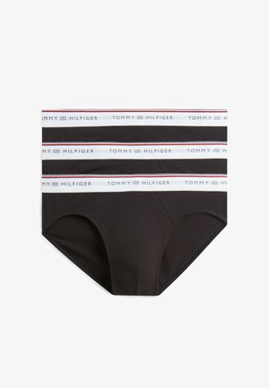 Trzy czarne męskie slipy z białymi elastycznymi paskami w talii, każdy z napisem "Tommy Hilfiger" i małym logo flagi w kolorach niebieskim, czerwonym i białym.