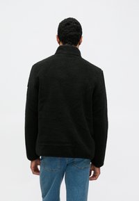 Fekete polárdzseki magas gallérral, texturált anyagból és laza szabással, világoskék farmernadrág fölött viselve. A háttéri nézet további részleteket nem mutat.