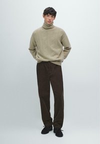 Hellbeiger, grob gestrickter Rollkragenpullover kombiniert mit dunkelbraunen Cordhosen mit lockerer Passform und feiner Struktur. Schwarze Schnürschuhe.