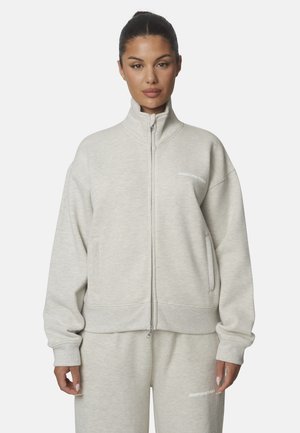 Donna con felpa leggera grigia con zip e pantaloni della tuta abbinati con piccolo testo bianco sul petto e sulla coscia, in piedi su sfondo bianco.