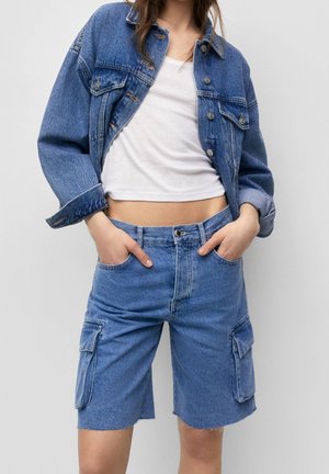 Person iført en blå denimjakke og cargoshorts med hænderne i lommerne og en hvid cropped top mod en ensfarvet baggrund.