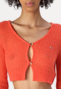 Pomarańczowy krótki kardigan z miękkiego, włochatego materiału, z głębokim dekoltem w kształcie litery V i zapięciem na guziki. Ozdobiony małym logo przy ramieniu.