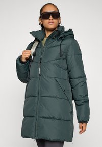 Abrigo tipo puffer en verde oscuro con capucha, que cuenta con un cierre de cremallera en el frente, dos bolsillos laterales y puños acanalados. El material parece acolchado y texturizado.