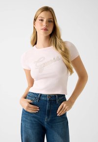 Light pink cropped t-shirt with ribbed texture and short sleeves, featuring a decorative silver logo. Paired with blue high-waisted jeans.