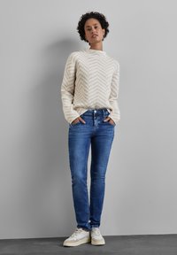 Knit sweater dengan pola chevron berwarna krem dan beige, dipadukan dengan celana jeans denim biru dan sepatu sneakers krem, berdiri di depan latar belakang abu-abu.