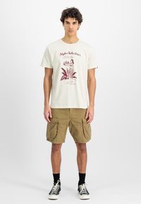 Beige puuvillane t-särk, millel on punane graafiline print naisest ja tekst, koos khaki värvi cargo lühikeste pükstega, millel on suured küljetaskud.