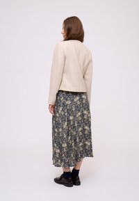 Donna che guarda di lato, indossando una giacca crema strutturata, una gonna midi a fiori navy con pattern, calze scure e scarpe oxford nere su sfondo bianco.