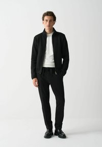 Melns ribots jaku ar rāvējslēdzēju virs balta apaļā kakla krekliņa, apvienots ar melniem šauriem biksēm un melniem auklējamiem apaviem. Minimalistisks dizains.