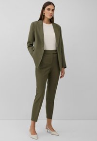 Completo sartorial verde oliva con giacca attillata e pantaloni a vita alta. Sotto, un top bianco testurizzato, completato da décolleté beige a punta.
