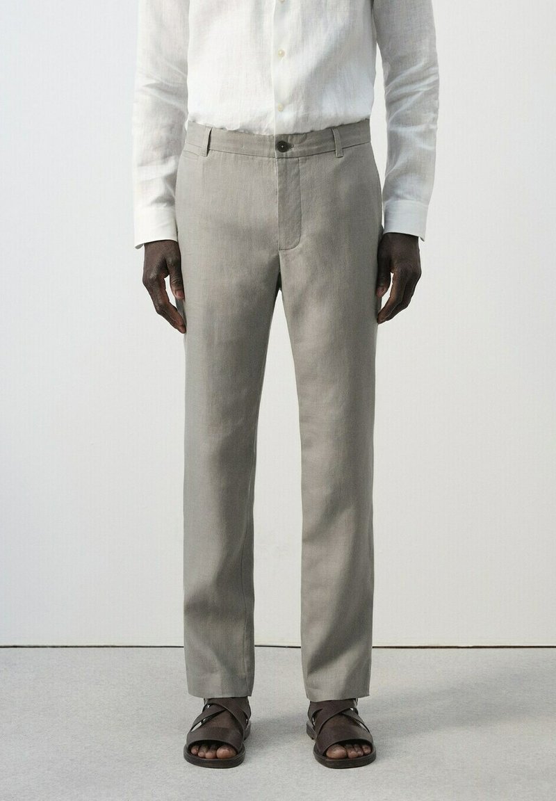 Homme portant un pantalon droit gris clair, une chemise blanche à manches longues boutonnée et des sandales marron foncé, debout sur un sol gris.