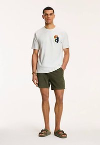 Weißes Baumwoll-T-Shirt mit einem mehrfarbigen, gestickten Logo auf der Brust, kombiniert mit dunkelgrünen Shorts und schwarzen Sandalen.