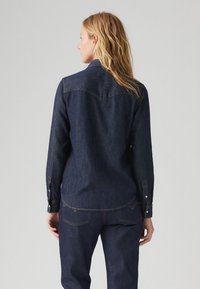 Camisa de ganga escura com mangas longas, punhos com botões e colarinho pontudo. Apresenta um yoke nas costas e detalhes de costura subtis.