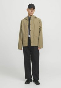 Khaki regenjacket met een capuchon, drukknoopsluiting en twee voork pocket, gedragen over een wit shirt en zwarte broek, gecombineerd met zwarte schoenen en een pet.