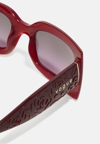 Occhiali da sole rossi con un design massiccio, dotati di lenti sfumate e intricati motivi floreali in rilievo sulle tempie. Presenti accenti dorati con il logo.