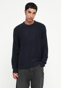 Pull en tricot bleu marine avec un col rond, des poignets côtelés et un tissu texturé. Porté avec un jean gris foncé, avec des détails de design minimalistes.