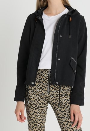 Femme portant une veste noire à capuche, un t-shirt blanc et un pantalon imprimé léopard avec fermeture éclair et détail anneau.