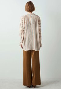 Striped long-sleeve shirt in cream and rust, with a loose fit and buttoned back detail. Paired with high-waisted, wide brown trousers.