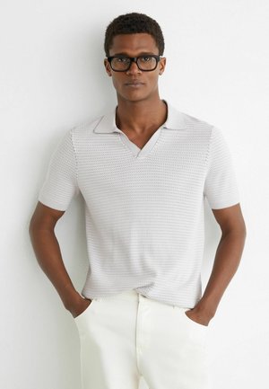 Férfi fekete szemüvegben, strukturált rövid ujjú fehér pólóingben és törtfehér nadrágban, zsebében tartva a kezét, fehér falnak támaszkodva áll.