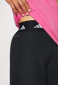 Leggings neri con una texture liscia e una vita alta, caratterizzati da accentuazioni del logo bianco. Una maglietta rosa a pois copre parzialmente la vita.