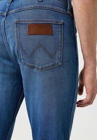 Jeans in denim blu con un'etichetta in pelle marrone Wrangler sulla tasca posteriore, caratterizzati da cuciture gialle e un design classico a gamba dritta.