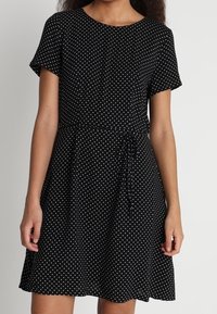 Robe noire à manches courtes avec des pois blancs, dotée d'un col rond et d'une ceinture ajustable à la taille pour une silhouette cintrée.