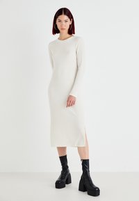 Vestido branco de manga longa e canelado com decote redondo e fenda lateral, combinado com botas de tornozelo pretas de plataforma. Textura suave, design minimalista.