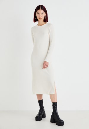 Rochie albă cu mânecă lungă, cu nervuri, guler rotund și fante laterale, asortată cu botine negre cu toc înalt. Textură fină, design minimal.