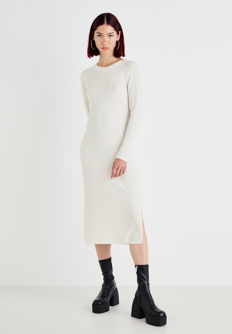 Vestido branco de manga longa e canelado com decote redondo e fenda lateral, combinado com botas de tornozelo pretas de plataforma. Textura suave, design minimalista.