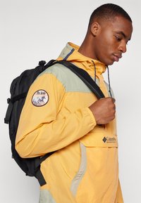 Gul og grøn vindjakke med et logo-patch, der har en frontlomme og justerbare snore, parret med en sort rygsæk.