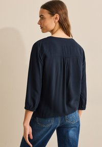 Marineblauw blouse met ¾ mouwen, voorzien van een geplooide rugdetail en een soepele, lichte stof, gecombineerd met blauwe denim jeans.