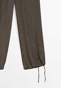 Pantalons en tissu vert olive foncé présentant un motif de feuilles subtil, des poignets élastiques et un détail de cordon au niveau de la cheville.