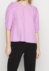 Blusa lilla con maniche corte a sbuffo e dettagli plissettati sulle spalle. Tessuto morbido, scollatura rotonda e vestibilità comoda. Abbinata a pantaloni neri.