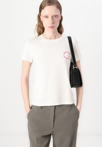 Hvid bomulds T-shirt med korte ærmer, rund halsudskæring og et rød/blå cirkulært logo på venstre bryst. Båret sammen med grå bukser og en sort taske.