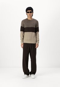 Maglione a maglia beige e marrone scuro con texture a coste, scollo rotondo e maniche lunghe; abbinato a pantaloni larghi marroni e sneaker bianche.