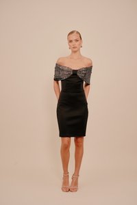 Sieviete stāv uz bēša fona, ģērbusies melnā kleitā ar atvērtām plecu daļām un teksturētu šalli, kas akcentētām bēša krāsas potīšu siksnām.