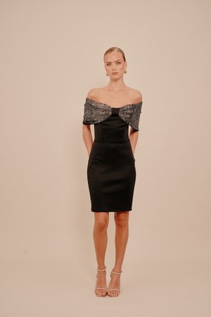 Vrouw staat tegen een beige achtergrond en draagt een zwarte off-shoulder jurk met een textuurdetail op de sjaal en beige enkelbandhakken.