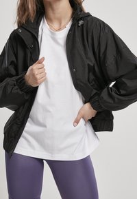 Zwarte lichtgewicht jas met een ritssluiting, elastische manchetten en een capuchon, gedragen over een wit T-shirt en paarse leggings. Gladde textuur.