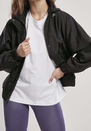 Černá lehká bunda se zapínáním na zip, elastickými manžetami a kapucí, nošená přes bílý tričko a fialové legíny. Hladká textura.
