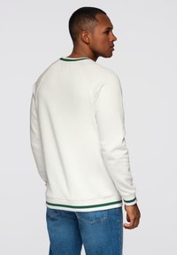 Hombre vestido con sudadera blanca con ribete verde y jeans azules, de pie y mirando a la derecha sobre un fondo claro y simple.