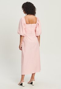 Vaaleanpunainen midi-mekko, jossa on neliömainen pääntie, puffihihat ja rypytetty yksityiskohta takana. Sujuva kangas ja lentävä siluetti.