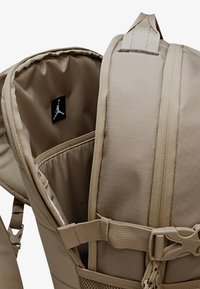Beigefarbener multifunktionaler Rucksack mit glattem Innenfutter, Reißverschlusstaschen und einem Stoff-Logoaufnäher, ausgestattet mit verstellbaren Riemen und robusten Beschlägen.