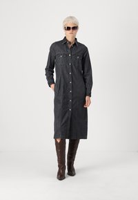 Čierna deninová dlhá šaty s gombíkovým zapínaním, dvoma náprsníkmi, dlhými rukávmi a rovnom strihom; skombinované s hnedými čižmami po kolená.