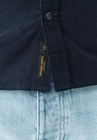 Marineblå kordfløjls skjorte med tekstureret overflade, knaplukning og en grøn label med teksten "DURABLE GOODS" i gult. Båret over lyseblå jeans.