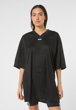 Ung kvinde iført en oversized sort perforeret FILA sportstrøje og sorte shorts, stående mod en ensartet lys baggrund.