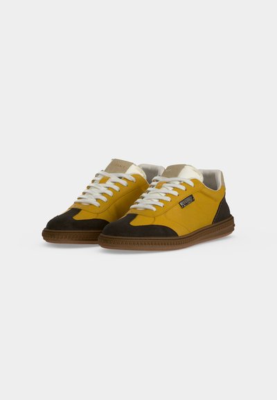 Paire de baskets basses avec des panneaux jaunes et noirs, des lacets blancs, une languette beige et des semelles en caoutchouc marron sur un fond uni.